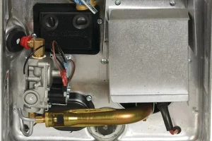 Close-up of a metal water heater component, showing valves, wiring, connections, and a protective cover. The setup appears complex and utilitarian.