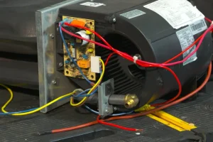 A black HVAC blower motor with attached wiring and circuit board, set on a wooden surface. Red, blue, and yellow wires create a complex, technical scene.