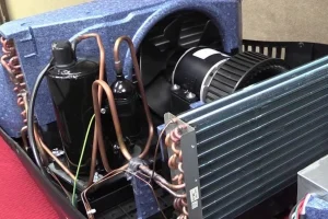 Close-up of an air conditioning unit's internal components. Visible are copper pipes, a fan, and a motor, conveying a technical and functional tone.
