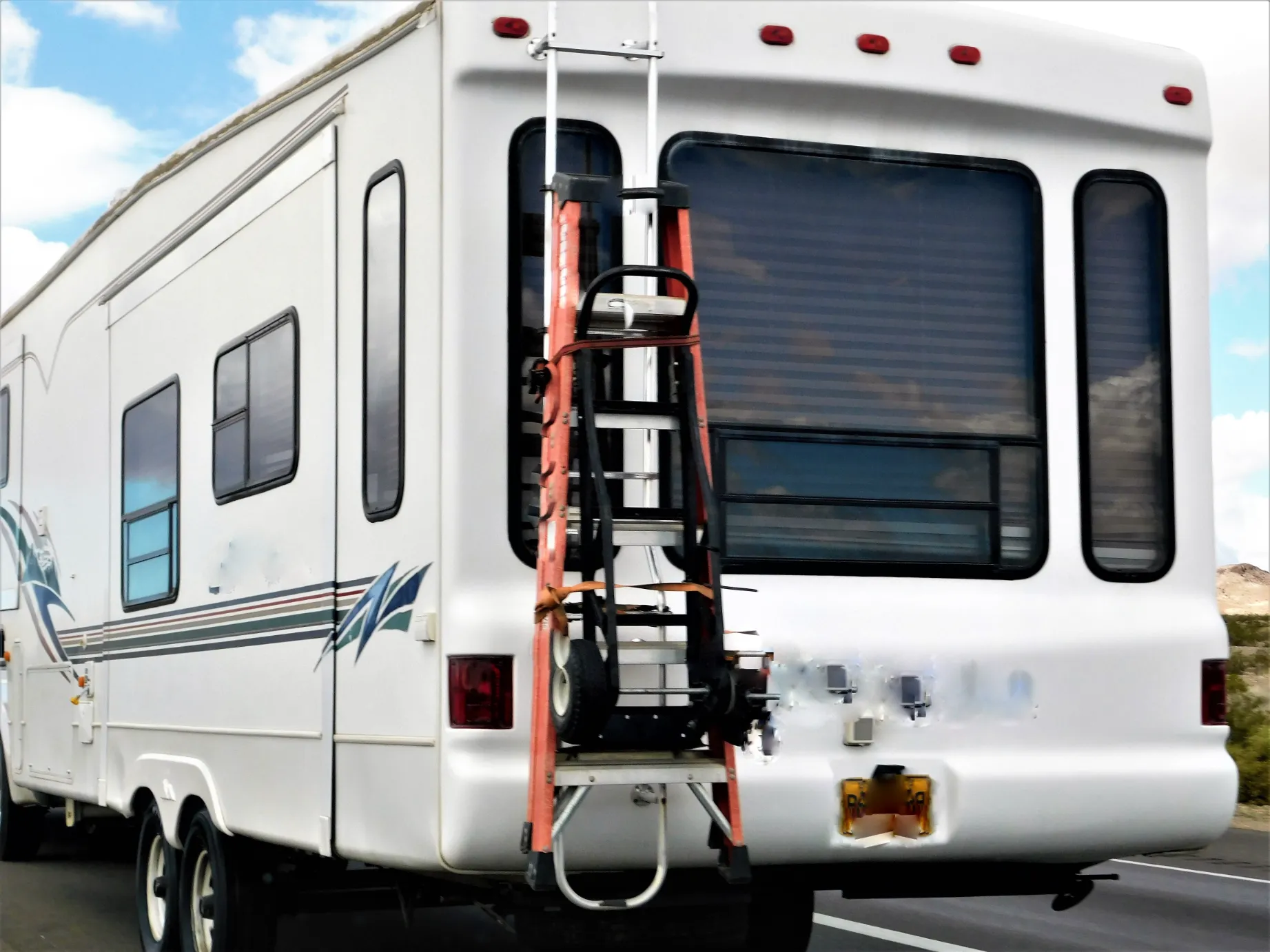 Rear view of a white RV on a highway. A red ladder and wheels are attached to the back. The sky is partly cloudy. The setting feels adventurous.